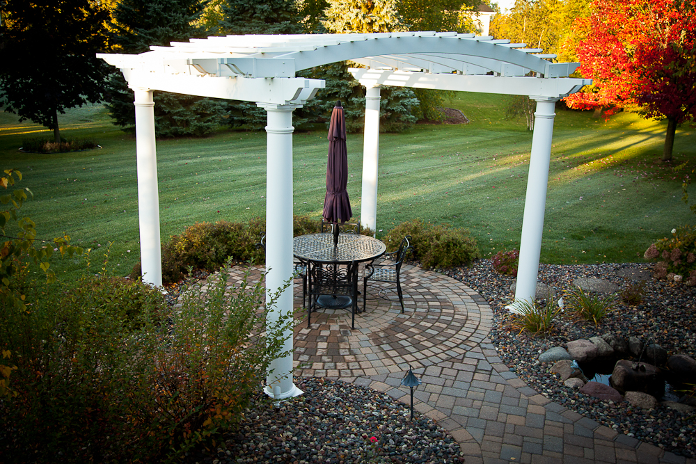 A white pergola with a round table, four chairs, and a closed umbrella stands on a curved stone patio surrounded by landscaping, grass, trees, and colorful autumn foliage.