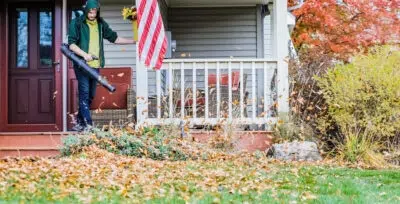 A person in a green hoodie uses a leaf blower to clear fallen leaves from a porch in front of a house with an American flag, autumn-colored trees, and well-maintained landscaping in the background.