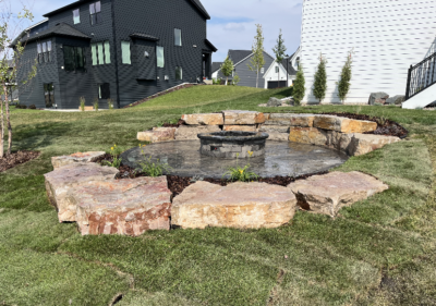 A stone fire pit with a circular rock seating area is set in a newly landscaped backyard with green grass and modern houses in the background.