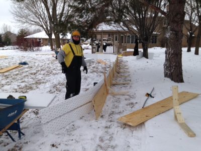 A person in winter clothing stands outdoors in a snowy yard, working on a partially built wooden structure with boards and tools scattered around—possibly preparing for snow removal. Trees and houses are visible in the background.