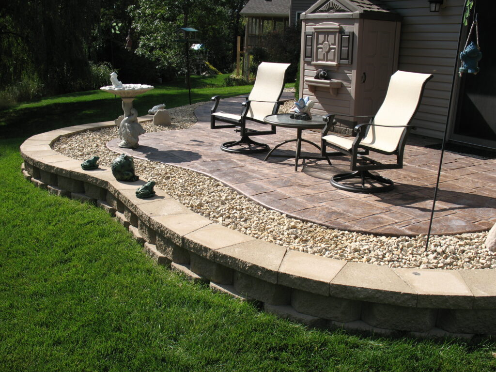 A backyard patio with two chairs and a table on stamped concrete, bordered by decorative stones and a low stone wall, overlooks a green lawn. A birdbath and a garden shed are also visible.