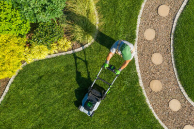 An aerial view of a person in a green hat mowing a vibrant green lawn with a cordless mower, bordered by colorful shrubs and a gravel path—perfectly maintained with help from local yard services.